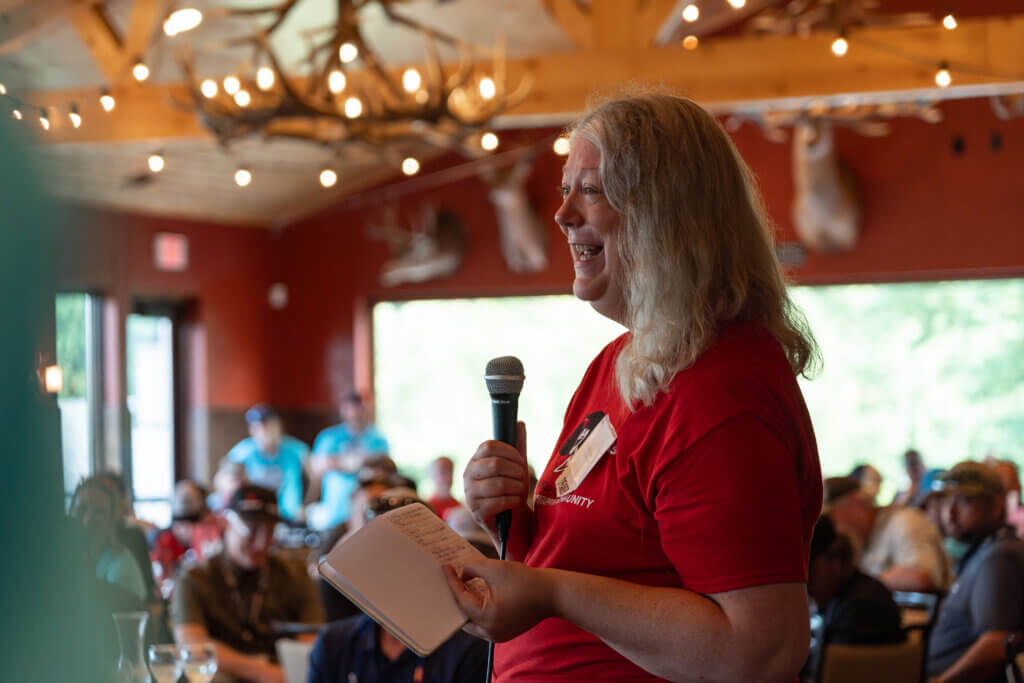 A person stands holding a microphone and a piece of paper in a room full of people.