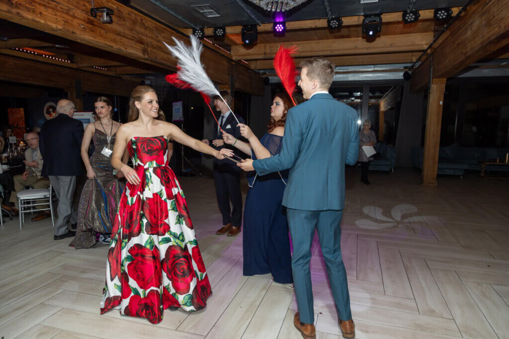 People dance together on a dance floor, one of them wearing a gown with red flowers on it and the otehr wearing a blue-gray suit.