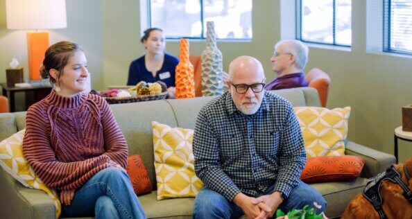 Two people sit on a couch. In the background, two people sitting on armchairs converse.