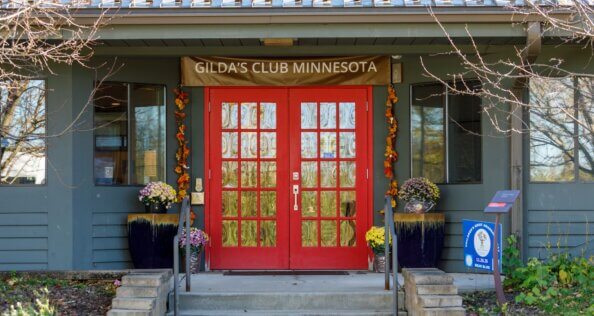 A gray-green building has red doors under a banner with "Gilda's Club Minnesota" printed on it.